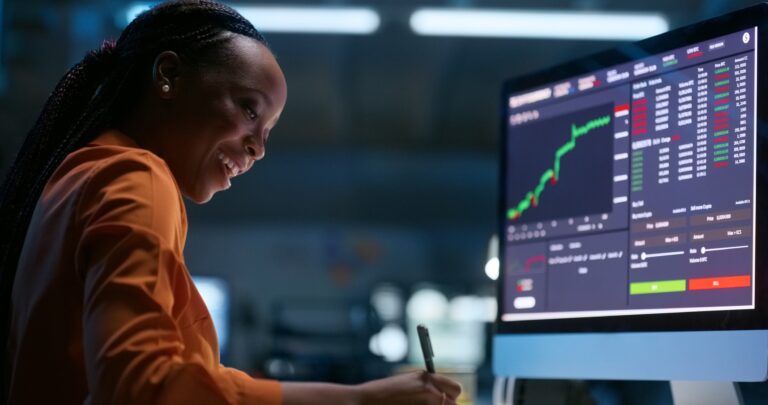 a person taking notes and smiling looking at stock charts on a computer