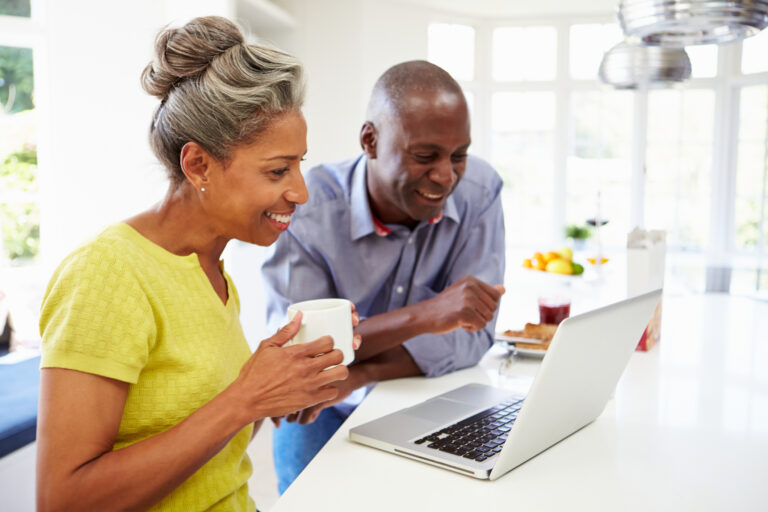 mature man and woman sitting in kitchen looking at laptop computer and smiling couple poc